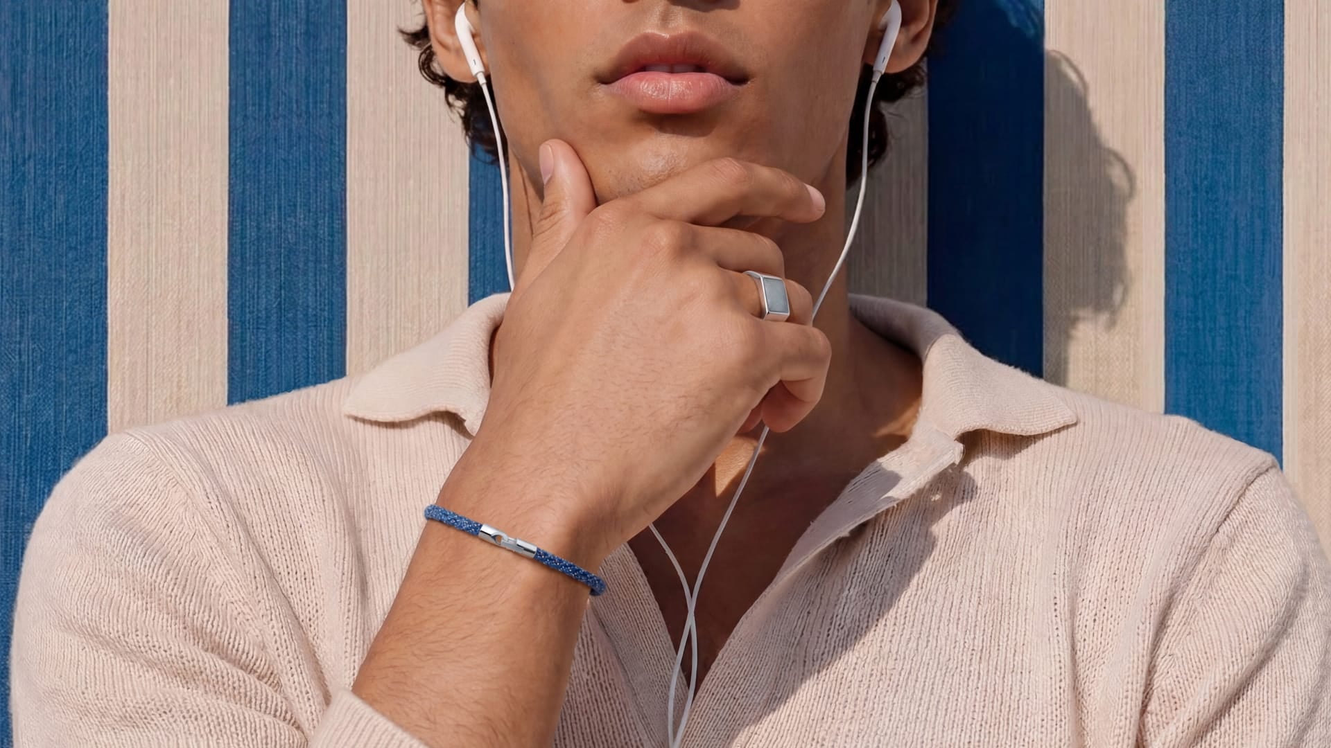 A close-up lifestyle shot of a man wearing a light beige knit polo shirt, white wired earphones, a blue beaded bracelet on his wrist, and a silver ring on his finger, set against a blue and cream striped background.