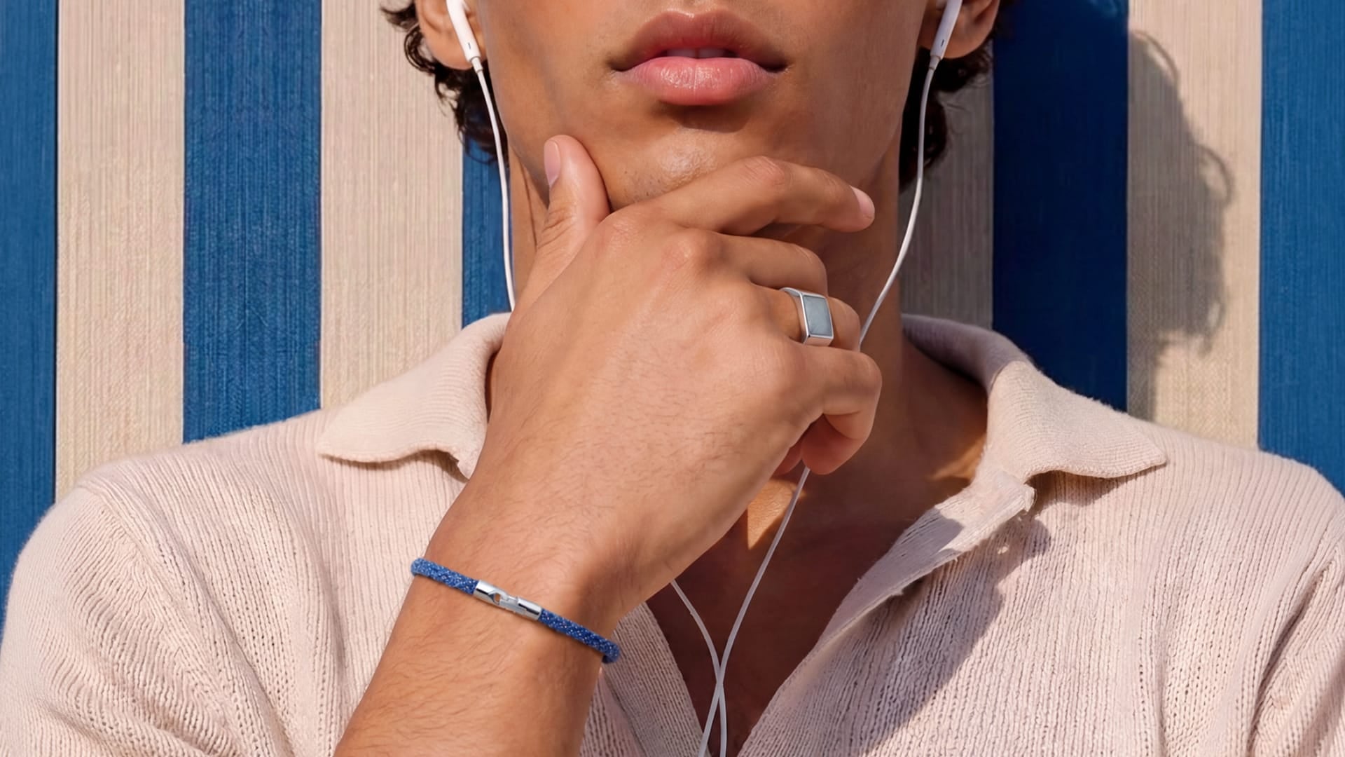 A close-up lifestyle shot of a man wearing a blue braided cord bracelet with a silver-toned metal band, along with white wired earphones and a silver ring, set against a blue and beige striped background.