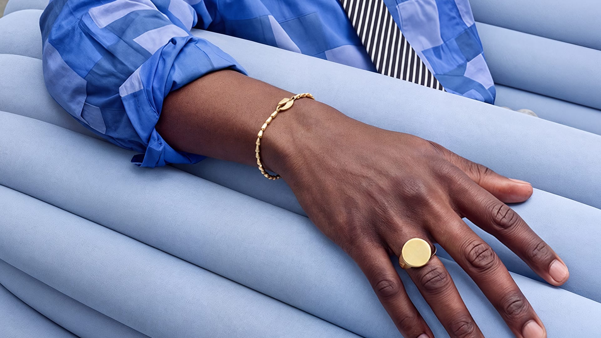 A close-up of a person's hand resting on light blue ribbed cushions, wearing a delicate gold chain bracelet and a large round gold signet ring.