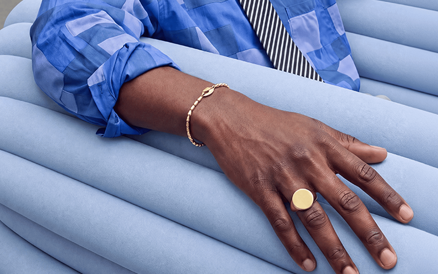 A close-up lifestyle shot of a person's hand resting on light blue rolled fabric, wearing a gold signet ring and a gold beaded bracelet. The person is also wearing a blue patterned shirt and a striped tie.