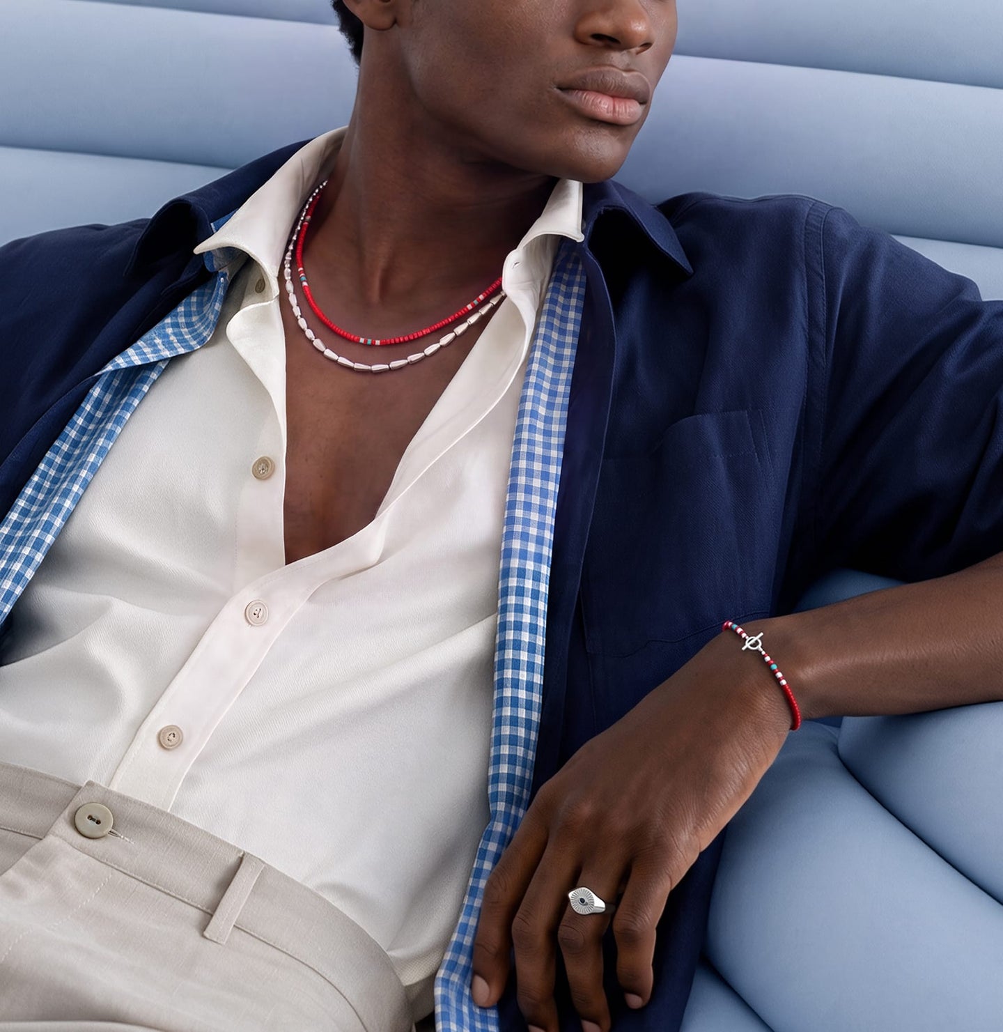 A close-up lifestyle shot of a model wearing a red and white beaded bracelet on one wrist and a matching red and white beaded necklace on the neck, styled with a blue and white gingham shirt, a navy blue overshirt, and beige trousers, seated on a light blue sofa.