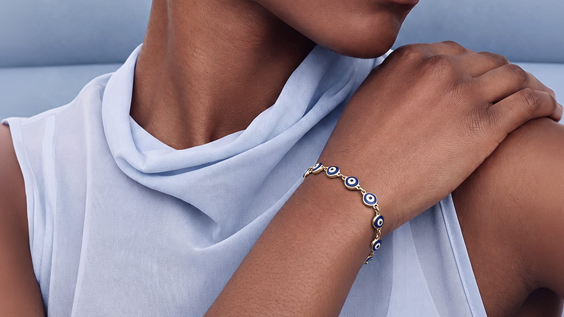 A close-up of a model wearing a delicate gold bracelet with blue and white evil eye charms, paired with a light blue draped top.