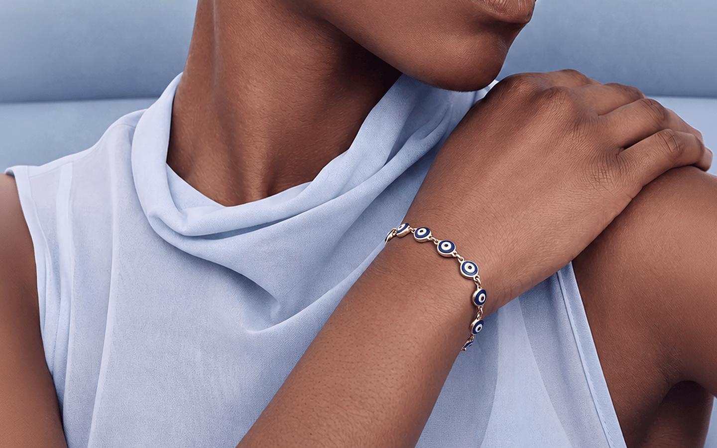 A close-up of a model wearing a delicate gold chain bracelet with blue and white evil eye charms, paired with a light blue draped top.