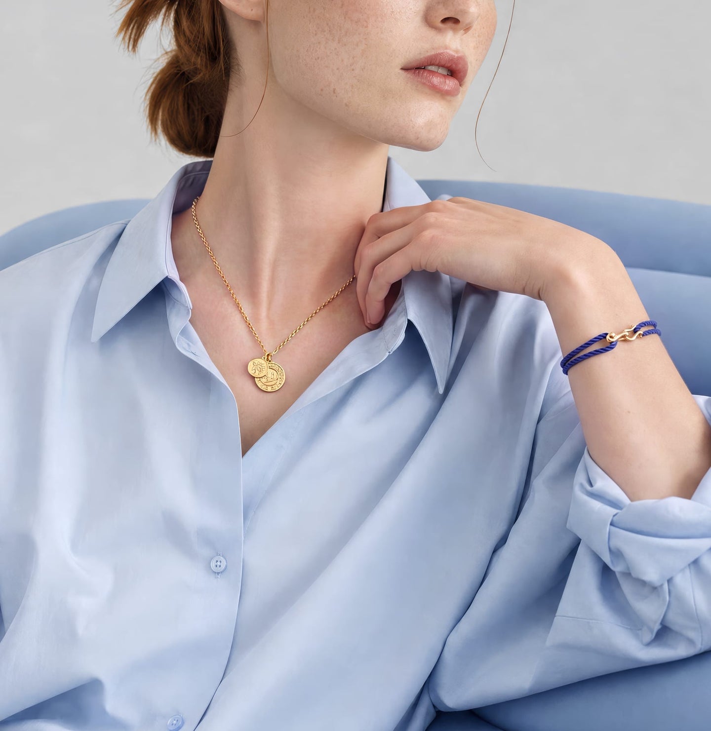 A close-up lifestyle shot of a model wearing a delicate gold necklace with a double-heart pendant and a matching gold bracelet with a blue stone accent, styled with a light blue button-down shirt.