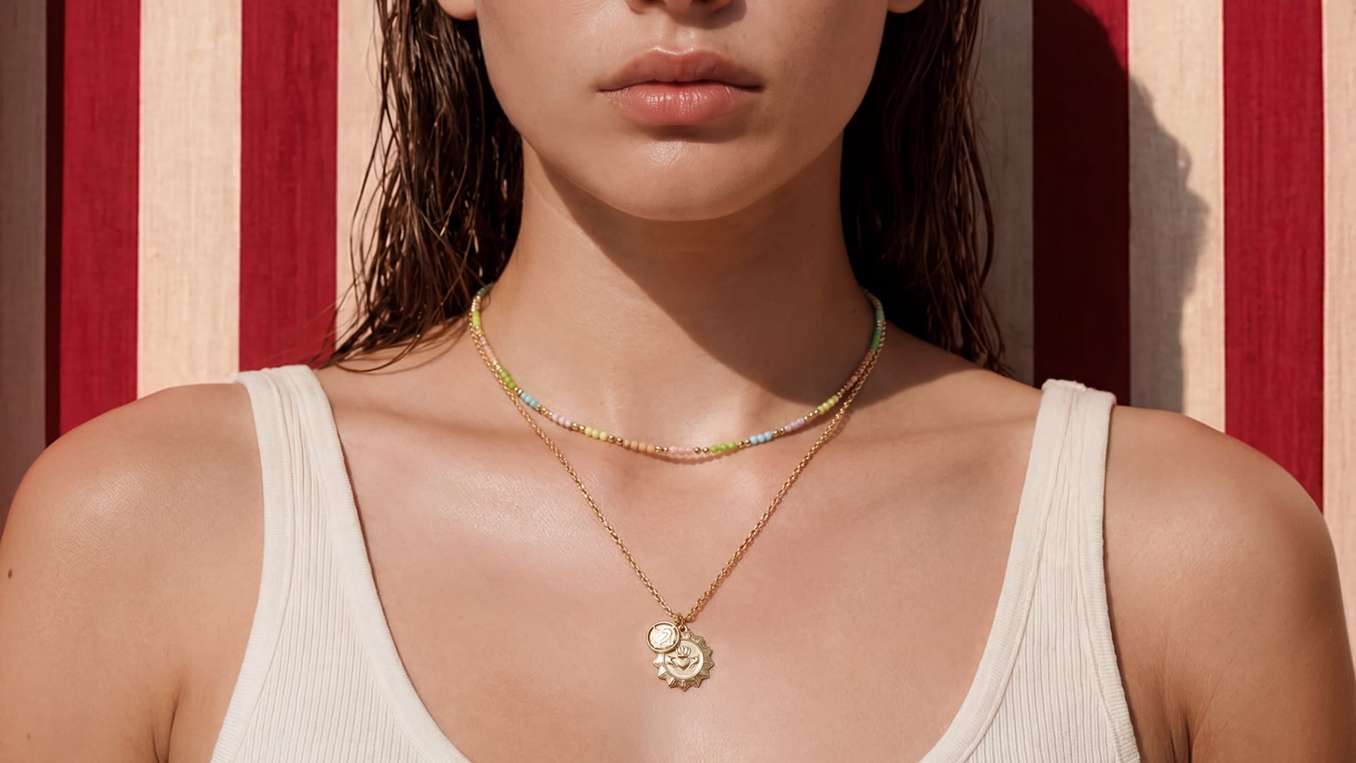 A close-up of a model wearing two delicate necklaces: a gold chain with a small sun pendant and a thin beaded choker necklace, set against a red and cream striped background.