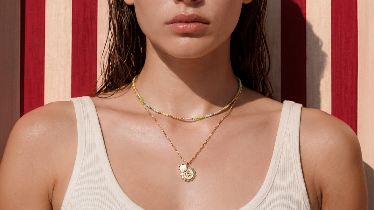 A close-up of a woman wearing layered necklaces, including a delicate gold chain with a circular pendant and a beaded choker, against a red and beige striped background.