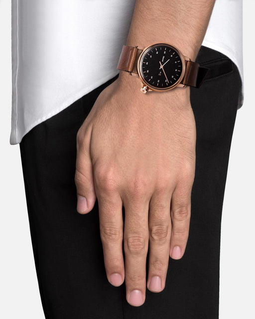 Miansai M12 Swiss watch with a rose tone stainless steel case, black dial, and cognac leather strap worn on the wrist